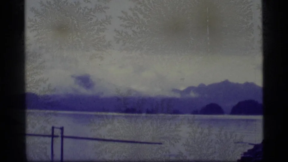 1977 Alaska 8mm frame showing calm lake water, distant mountain silhouettes, cloudy sky, and a foreground railing with aged-film artifacts.