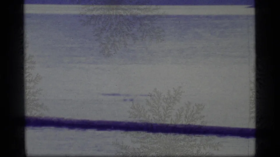 1977 Alaska 8mm frame with a pale lake surface, faint far shoreline, dark horizontal railing, and visible frost-like film artifacts.