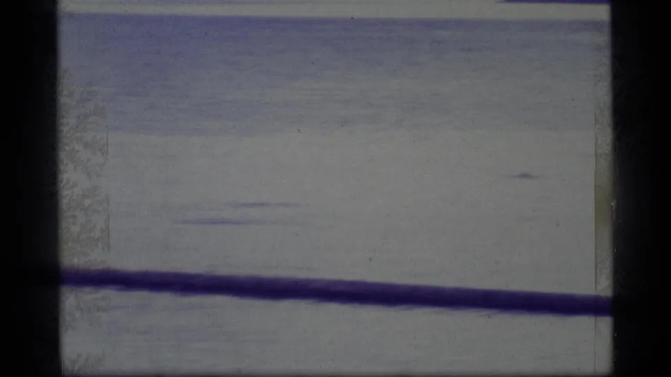1977 Alaska 8mm frame showing open lake water, a dark foreground railing, soft exposure, and scratches or frost-like emulsion texture.