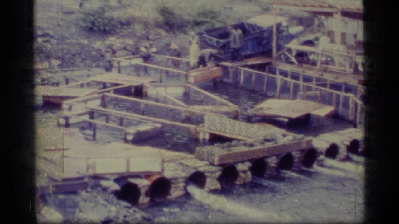 1977 Alaska 8mm frame showing an outdoor aquaculture setup with wooden raceways, water channels, pipes, and a worker near the tanks.