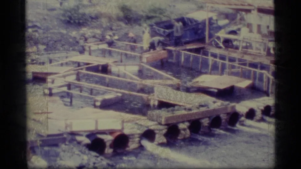1977 Alaska 8mm frame showing an outdoor aquaculture setup with wooden raceways, water channels, pipes, and a worker near the tanks.