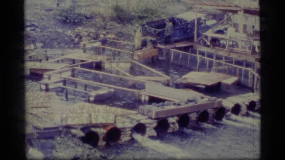 1977 Alaska home-movie frame of a freshwater fish facility with pipe sections, wooden raceways, water flow, and a distant worker.