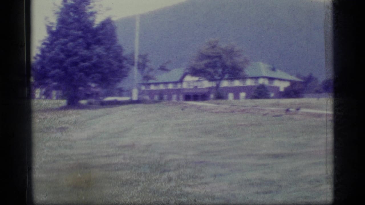 Duck on grass near long brown buildings with trees and hills behind.