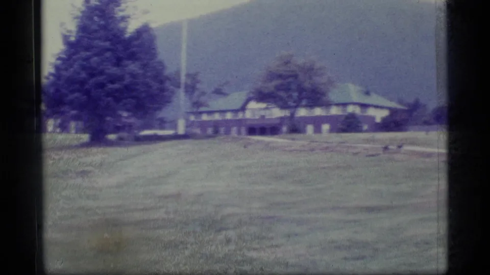 1977 Alaska 8mm frame showing a duck in a grassy field before low brown rural buildings and distant hills.