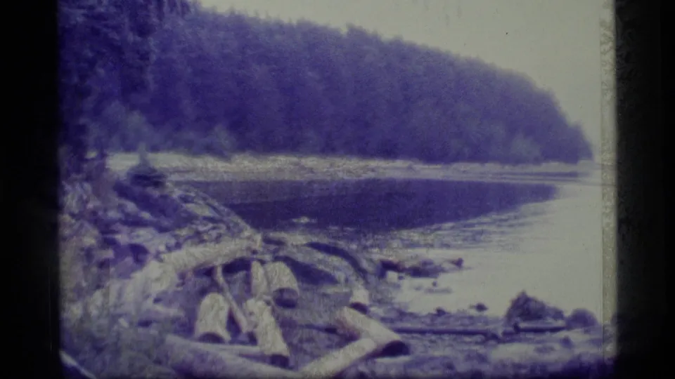 A wider 1977 Alaska home-movie view shows driftwood on the shore, still river water, and a dark forested slope across the bank.