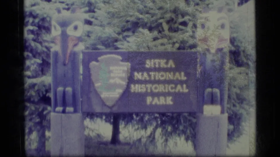 Close 1977 8mm view of the Sitka National Historical Park sign framed by carved totem forms and greenery.