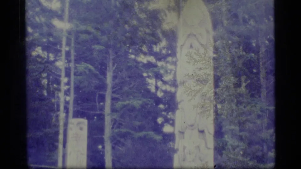 1977 8mm frame of a tall carved totem pole standing in the forest at Sitka National Historical Park.