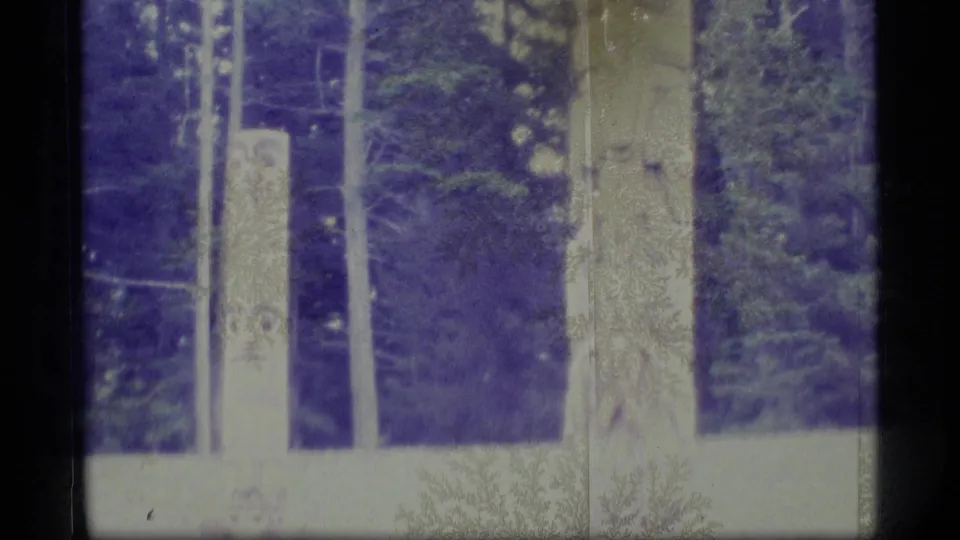 1977 Alaska home-movie frame of carved totem poles beside a low building, with evergreen trees in the background.