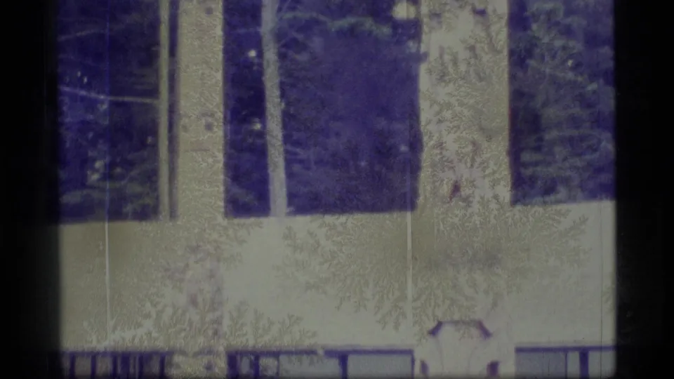 1977 Alaska 8mm frame showing the bases of carved totem poles near grass, dark railings, and part of a low building.