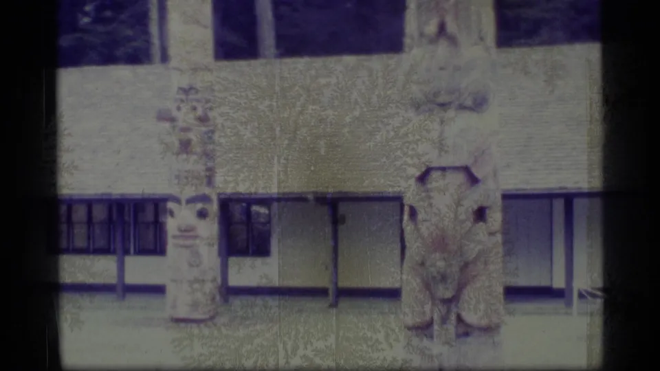 1977 Alaska home-movie frame with a carved figure or pole section beside railings, a low building, grass, and evergreen trees.
