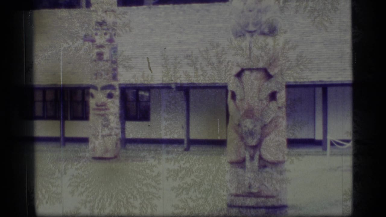 1977 Alaska 8mm frame showing two carved totem poles outside a long, low building with dark window panels.
