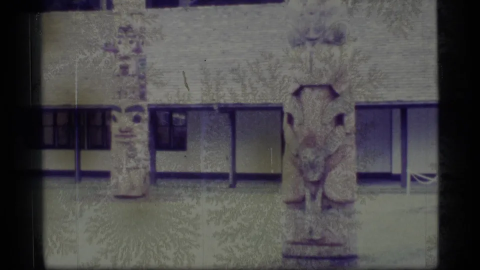 1977 Alaska 8mm frame showing two carved totem poles outside a long, low building with dark window panels.