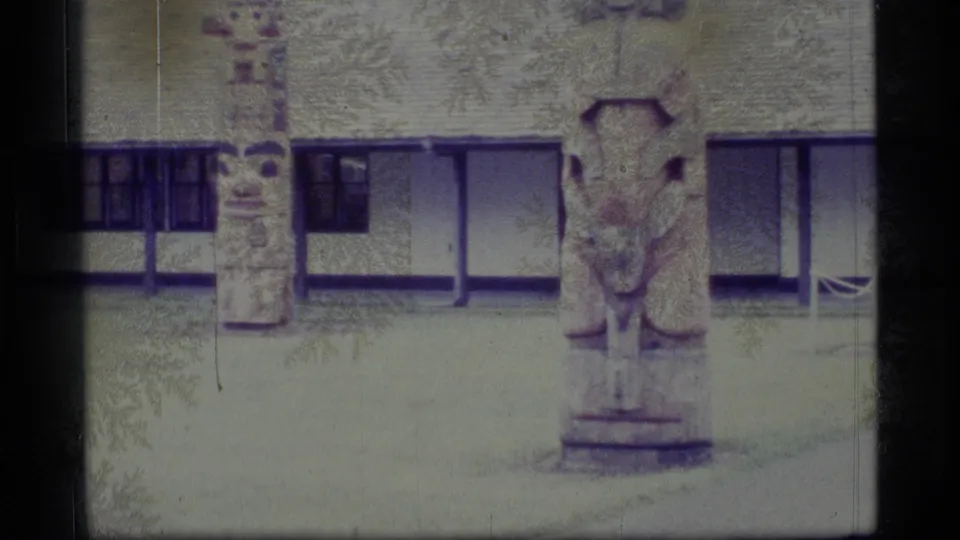 1977 Alaska home-movie view of carved totem poles beside a building, with soft grain and muted blue-green color.
