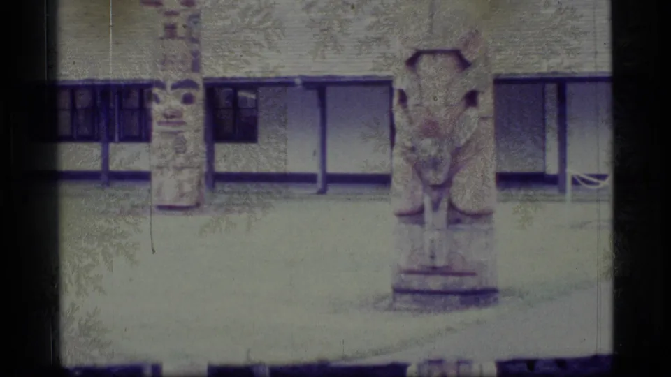 1977 Alaska 8mm frame with carved totem poles in front of a low building, recorded from a visitor viewpoint.