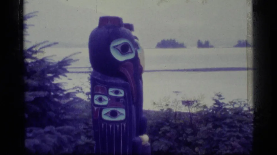 1977 Alaska frame showing a painted totem pole with bird-like features near a misty shoreline and distant trees.