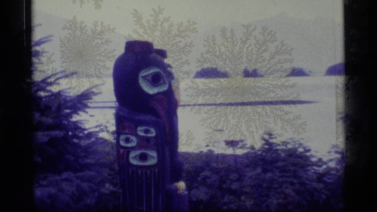 1977 Alaska 8mm frame of a painted totem pole in trees above a calm bay with low islands in the distance.
