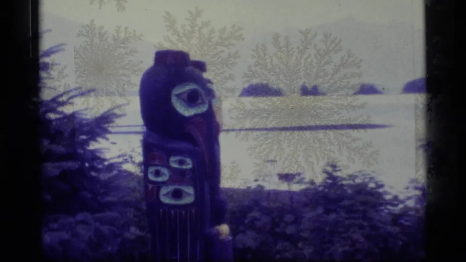 1977 Alaska 8mm frame of a painted totem pole in trees above a calm bay with low islands in the distance.