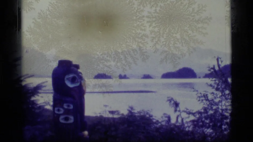 1977 Alaska home-movie frame showing a totem pole partly framed by trees, with water and small islands beyond.