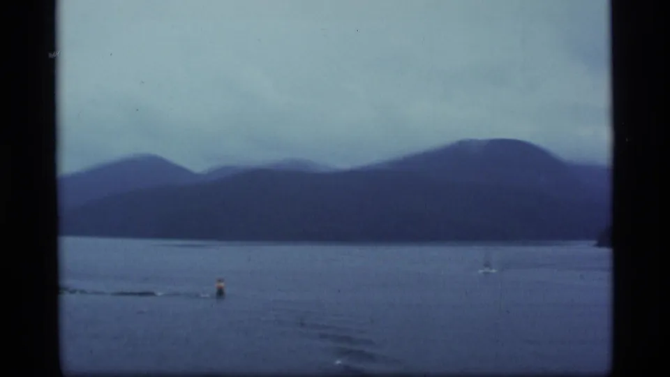 1977 Alaska 8mm frame looking across calm water toward blue-gray mountains, with a small boat visible on the bay.