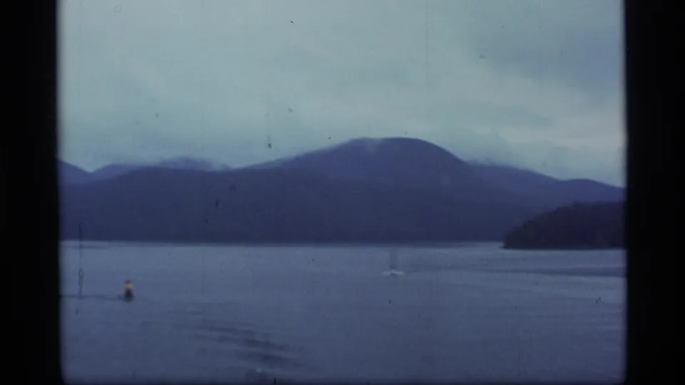 1977 Alaska 8mm frame showing a misty lake, layered mountains, a buoy, and a distant small boat under overcast skies.
