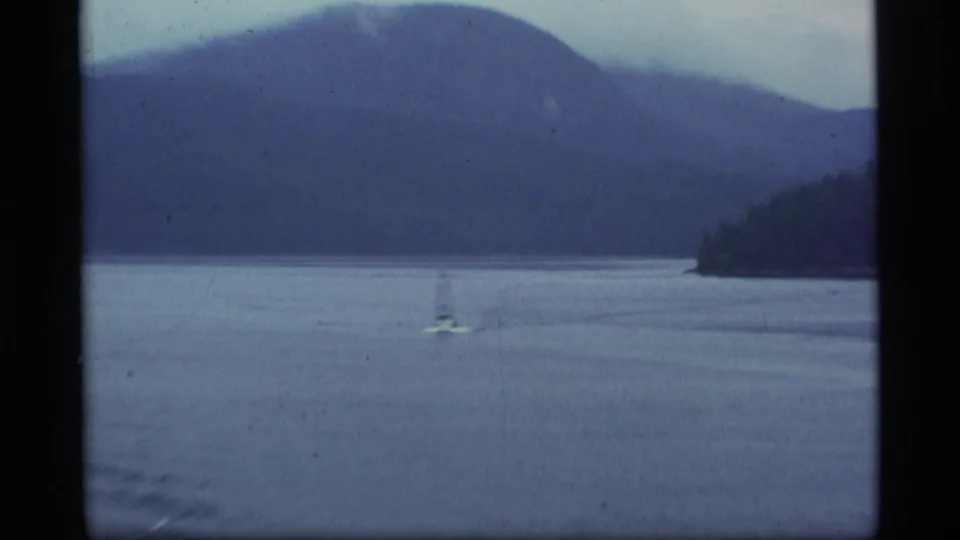 1977 Alaska 8mm frame of a small sailboat on calm lake water with wooded shoreline and misty mountains beyond.