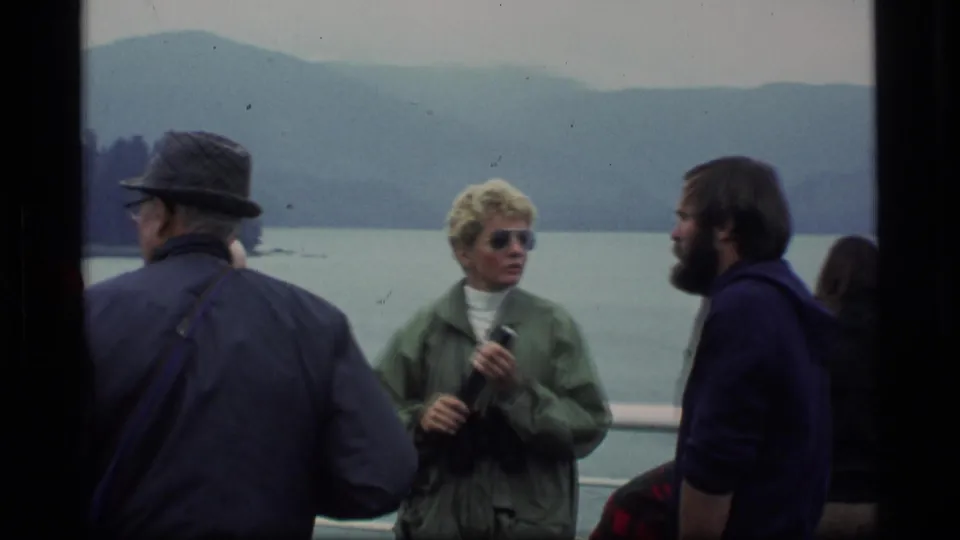 1977 Alaska 8mm frame showing adult passengers in jackets on a vessel deck, with lake water and mountains behind them.