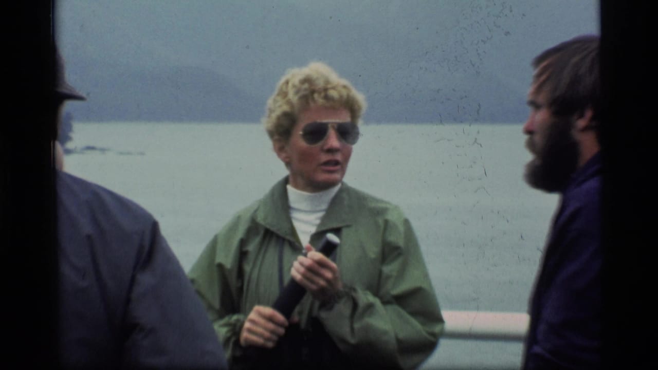 Woman in sunglasses holds a microphone on a ferry with men nearby and water behind her.