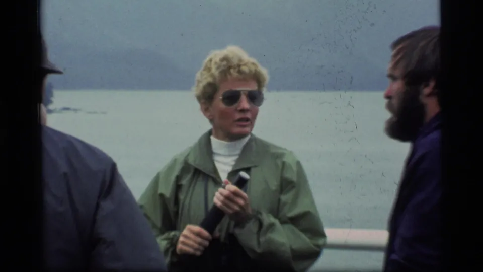In 1977 Alaska footage, a blonde woman in aviator sunglasses holds a microphone on a ferry while two men stand nearby.