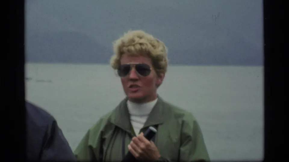 In the Alaska ferry scene, the woman faces the camera with a microphone, wearing sunglasses and a green jacket against gray water.