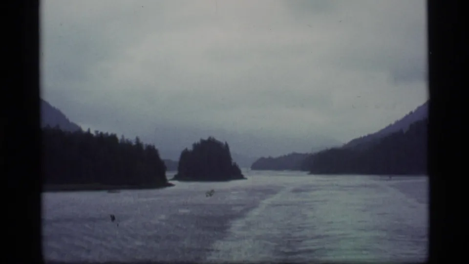 Wide 1977 Alaska 8mm view of a calm, misty water passage between forested islands beneath a gray overcast sky.