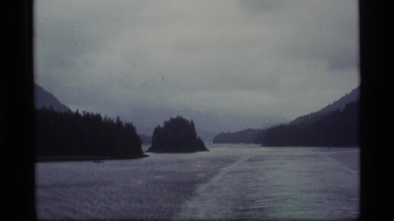 Boat view across a calm waterway toward wooded islands and dark hills under an overcast sky.