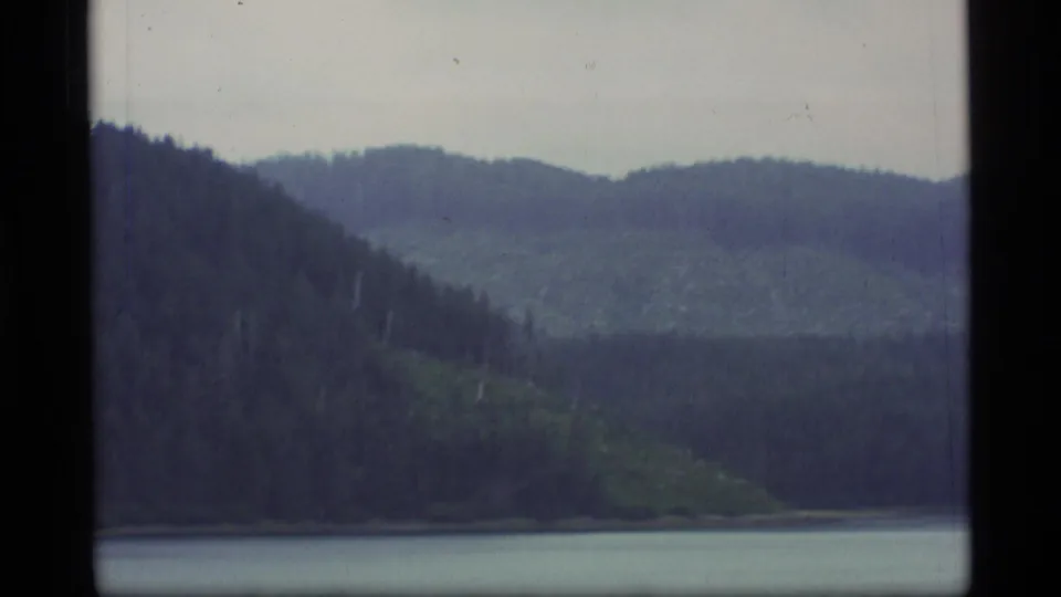 1977 Alaska 8mm frame of a forested shoreline, layered green hills, and still water seen from a boat.
