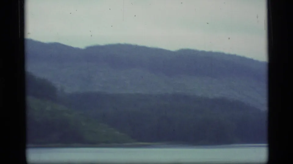 1977 Alaska 8mm boat-view frame with muted hills, evergreen slopes, and a calm waterline in soft focus.