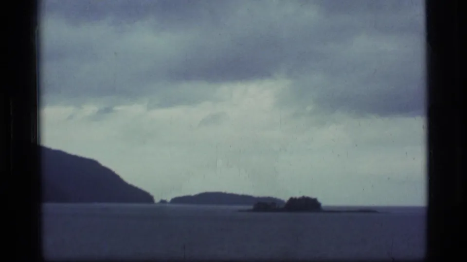 1977 8mm Alaska coastal view with open water, small low islands, and a dark forested headland below a cloudy sky.
