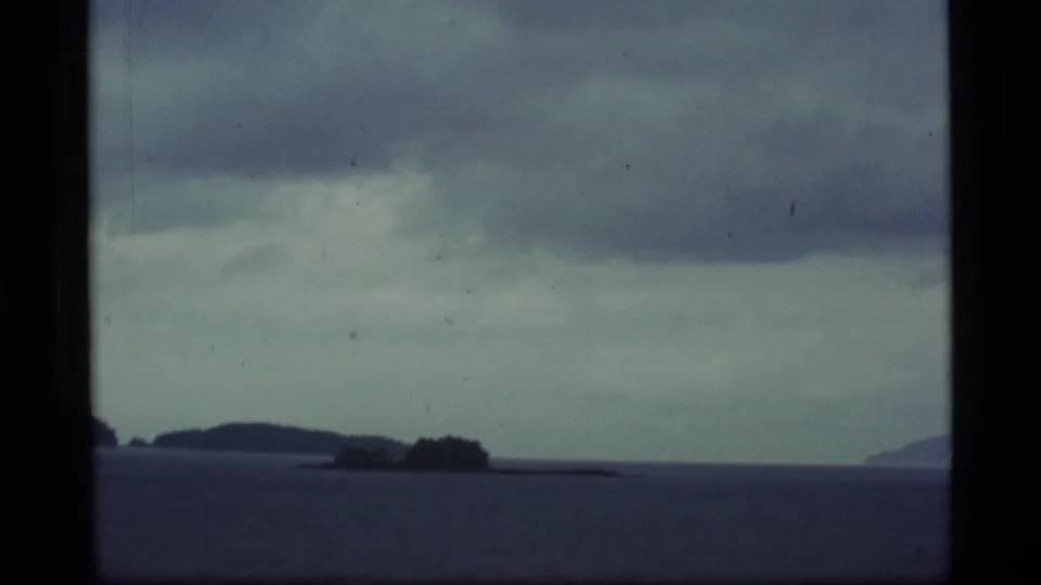 1977 8mm Alaska home-movie frame showing small islands across calm coastal water with distant shoreline shapes under overcast clouds.