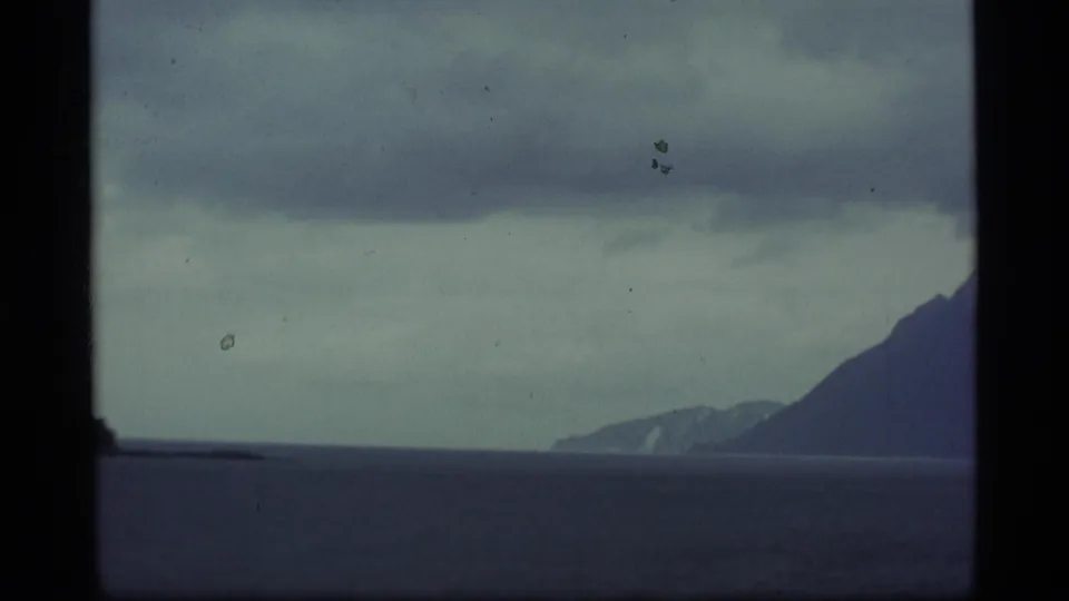 1977 8mm Alaska coastal landscape with broad water, distant mountain shoreline, heavy clouds, and a dark forested slope at the right edge.