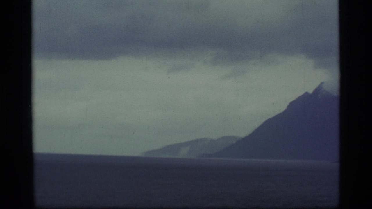 1977 Alaska 8mm frame showing a distant mountain ridge and dark water beneath low cloud, bordered by the film frame.