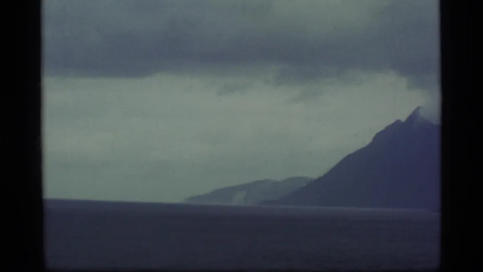 1977 Alaska 8mm frame showing a distant mountain ridge and dark water beneath low cloud, bordered by the film frame.