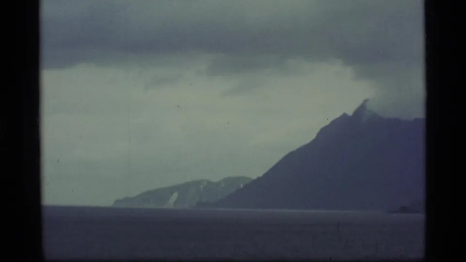 1977 Alaska home-movie frame with a steep volcano-shaped mountain mass rising beside open water under overcast skies.