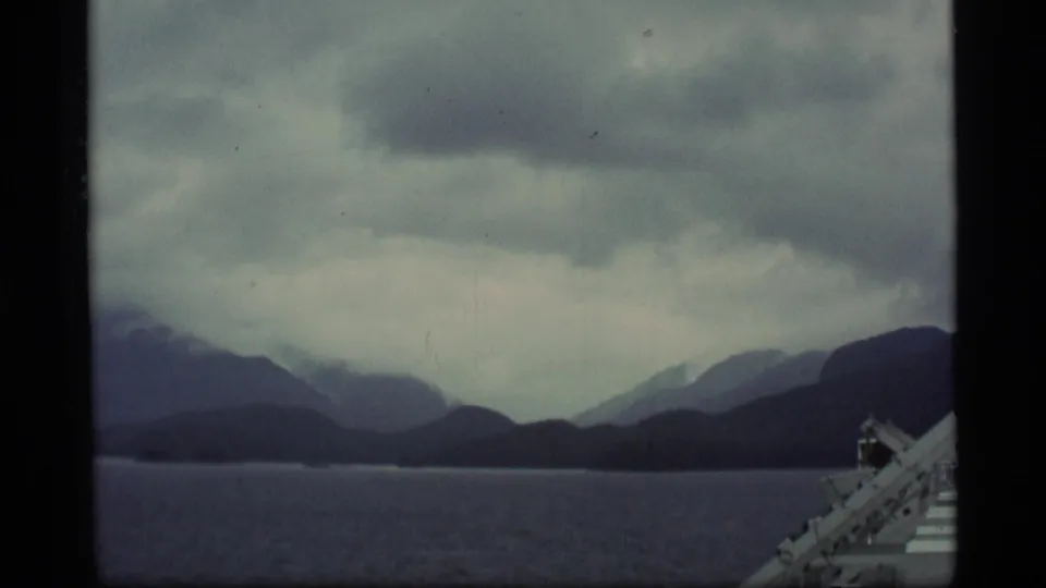 1977 Alaska home-movie frame of mist-softened mountains across dark water, with a small vessel or railing element at the lower right edge.