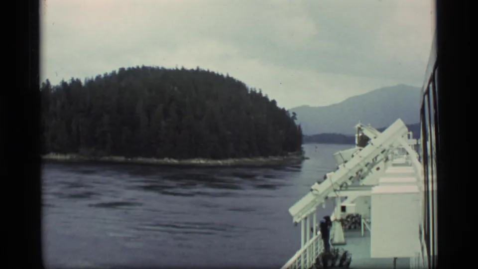 Alaska cruise view with a forested island across calm water, ship railings, and deck structure at the right edge.