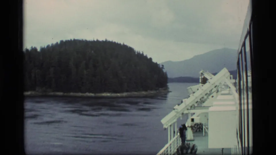 1977 Alaska home-movie frame showing ship deck equipment at right and a tree-covered island across the water.