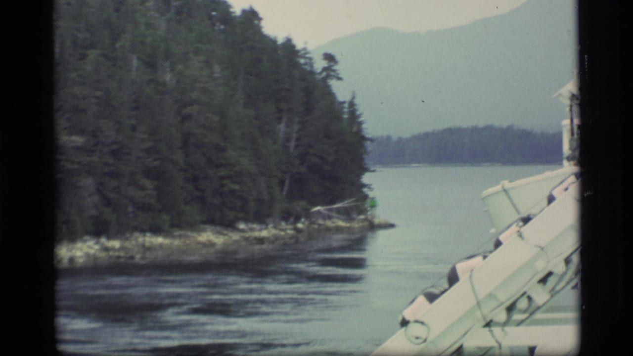 Ship rail beside a forested shoreline, calm water, and distant mountains.