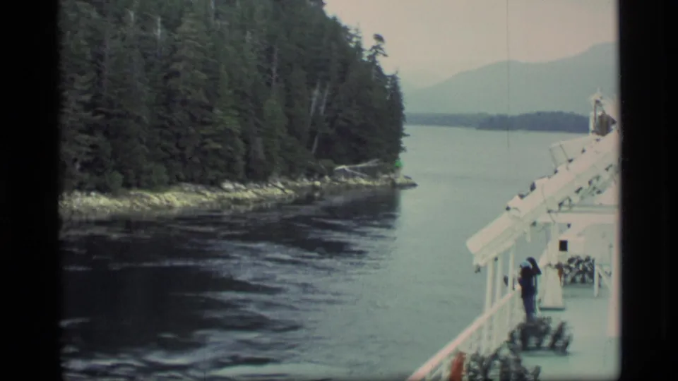 1977 Alaska shipboard view with a passenger on deck as the vessel moves past evergreen trees, rocky shore, and calm water.