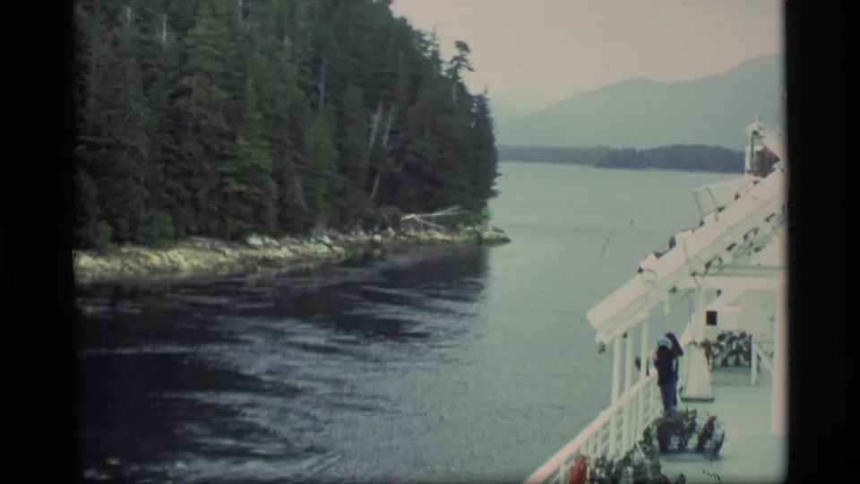 1977 Alaska 8mm frame showing a passenger on a ship deck, forested coast to the left, and muted mountains over the water.