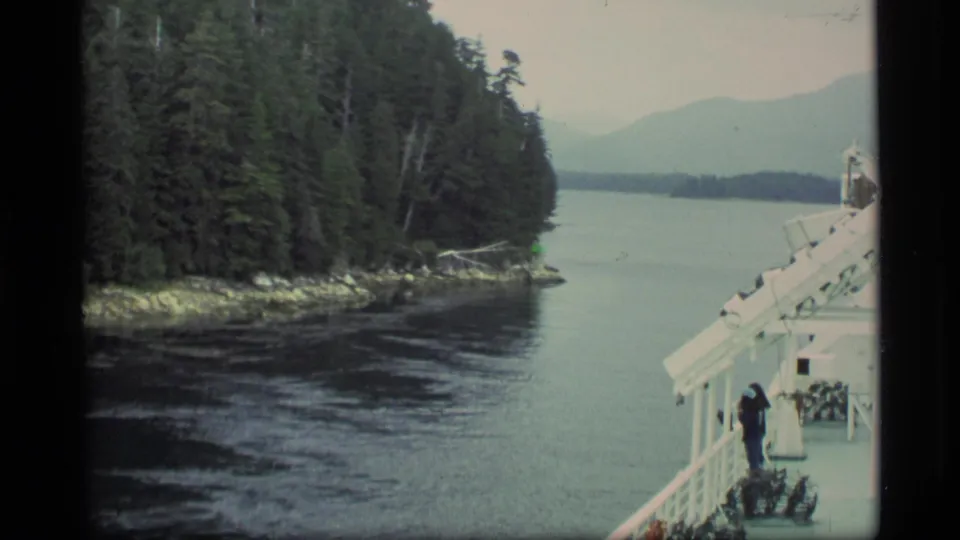 1977 Alaska home-movie frame from a moving vessel, with a passenger at the rail and dense trees along a narrow water passage.