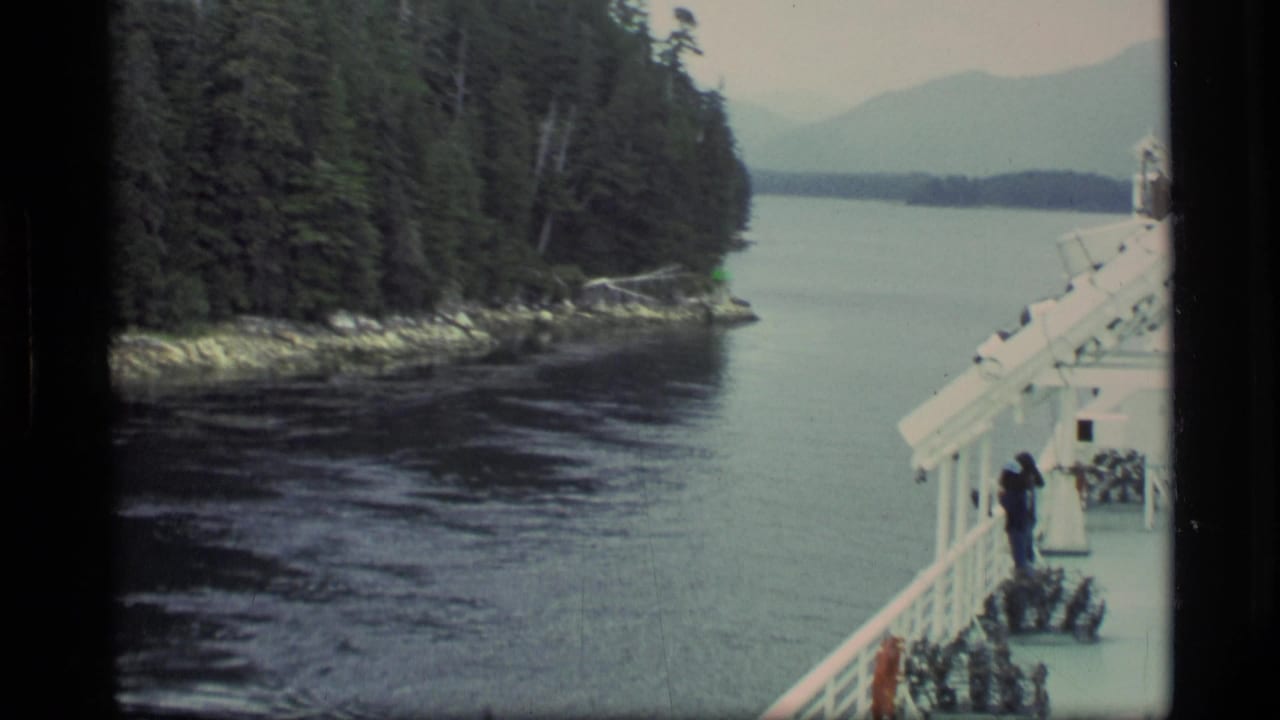 Boat deck passing a forested shoreline and narrow waterway with distant mountains.