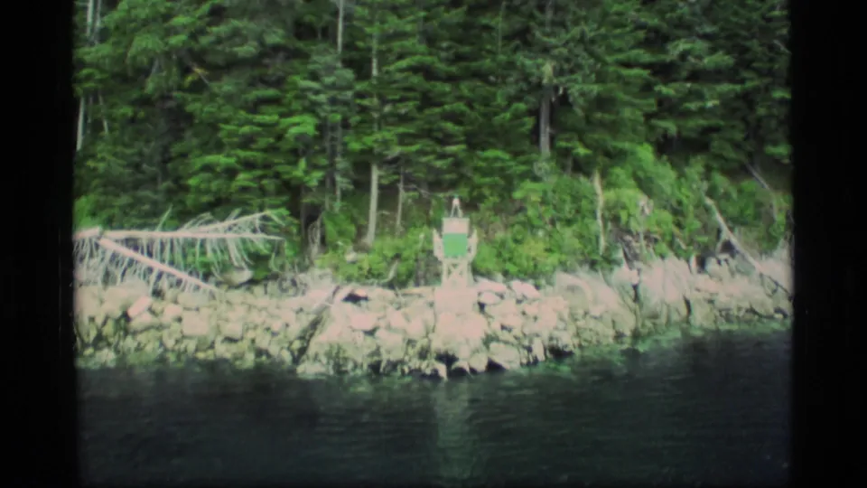 Closer 8mm view of an Alaskan rocky shoreline, dense evergreens, fallen wood, and a small green-and-white marker by the water.