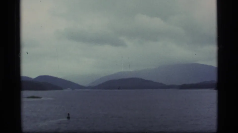 1977 Alaska 8mm landscape with open water, layered distant mountains, low islands, and heavy overcast sky.