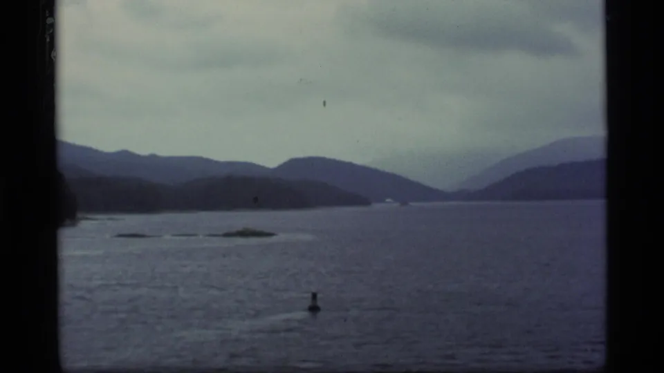 Alaska lake passage shows small water markers, low forested land, and hazy mountain ridges in faded 8mm color.